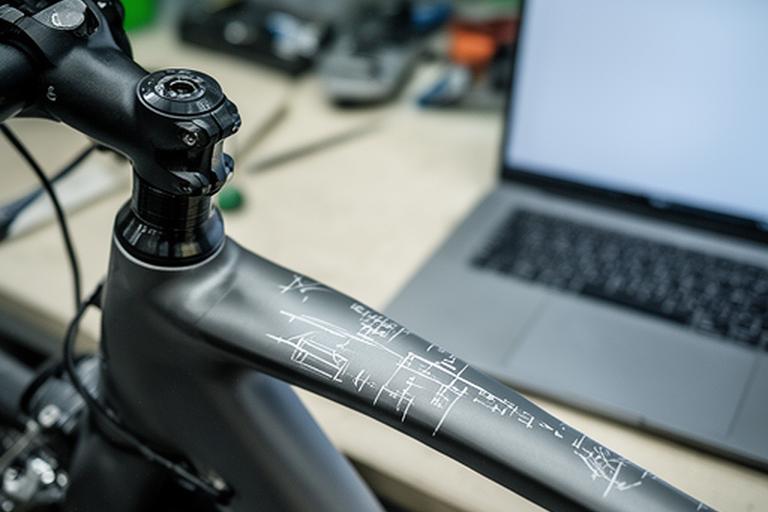 Detalhe de quadro de bicicleta com marcações de manutenção e um computador portátil em cima da bancada